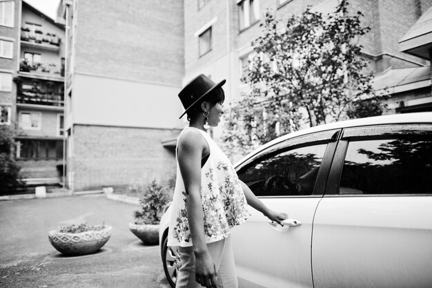 Incroyable femme modèle afro-américaine en pantalon vert et chapeau noir posé près de la porte blanche de la voiture