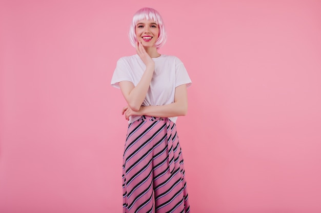 Incroyable jeune femme au Pérou à la mode touchant sa joue avec le sourire. Portrait intérieur d'une femme caucasienne raffinée en t-shirt blanc et perruque rose