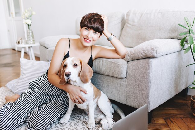 Incroyable jeune femme porte un pantalon rayé et montre-bracelet posant sur le sol tout en jouant avec un chien beagle