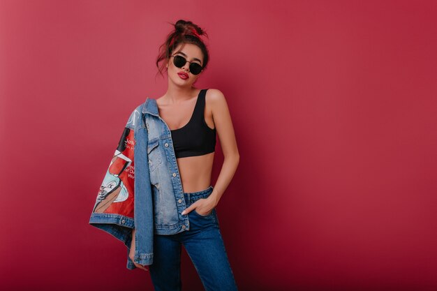 Incroyable modèle féminin avec une coiffure rétro debout dans une pose confiante et tenant une veste en jean