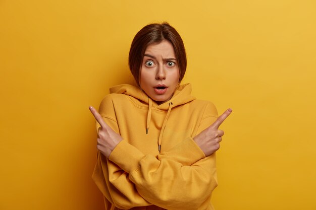 Indécis et perplexe jeune femme douteuse pointe du doigt sur le côté gauche et droite, hésite entre deux varinats ou produits, demande votre avis, a besoin de conseils au choix, porte un sweat-shirt jaune