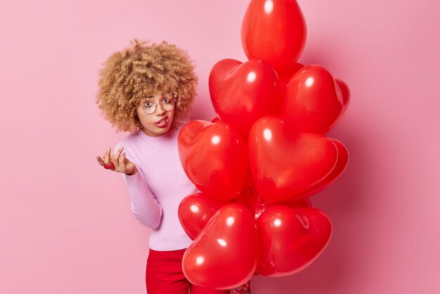 Indignée, frustrée, la femme bouclée hausse les épaules semble perplexe devant la caméra tient un gros tas de ballons cardiaques n'aime pas quelque chose que les lèvres ont une fuite de maquillage isolé sur fond rose