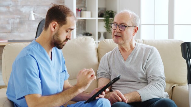Infirmier de race blanche parlant avec un patient d'une maison de retraite au sujet de sa santé. L'infirmière prend des notes sur une tablette numérique