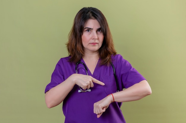 Infirmière d'âge moyen portant l'uniforme et avec un stéthoscope pointant sur sa main rappelant l'heure avec une expression sceptique sur le visage sur un mur vert isolé