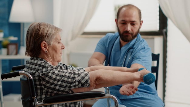 Infirmière aidant le patient handicapé avec des exercices physiques pour la récupération. Femme à la retraite assise dans un fauteuil roulant à l'aide d'haltères pour une activité de guérison dans une maison de soins infirmiers pendant que l'infirmière apporte son soutien