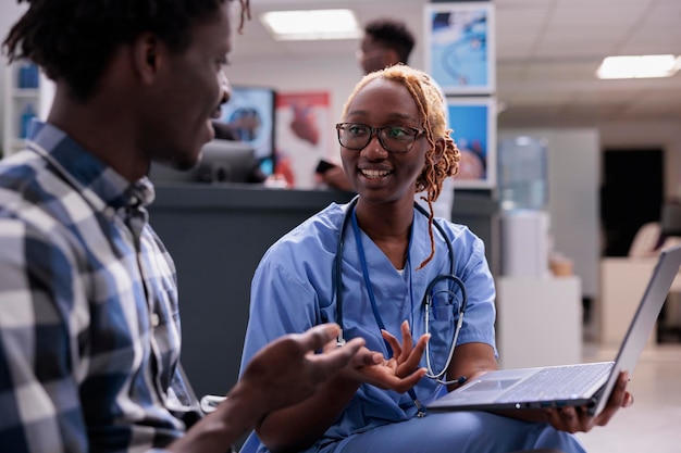 Infirmière consultant un patient à la clinique, assise dans le hall de la salle d'attente et utilisant un ordinateur portable. Expliquer le diagnostic pour donner un traitement de récupération avec une assurance maladie et un soutien, zone d'attente.