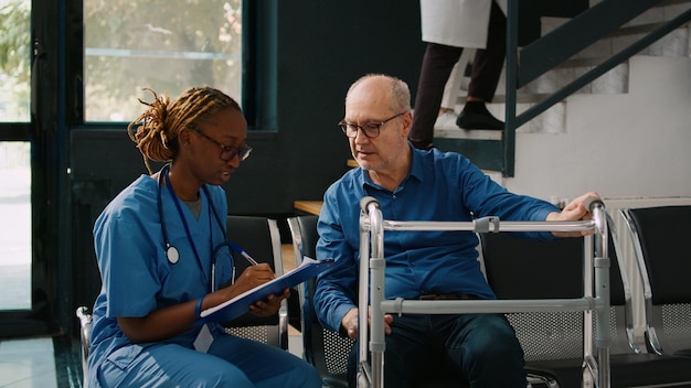 Infirmière faisant une consultation de contrôle avec un vieux patient dans la zone d'attente de l'hôpital, ayant un déambulateur. Assistant médical rédigeant un rapport clinique ou un formulaire de rendez-vous, centre de santé.