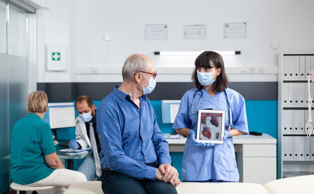 Infirmière et homme âgé avec des masques faciaux regardant une tablette avec une image de cardiologie à l'écran. assistant expliquant le diagnostic cardiovasculaire sur un gadget à un patient âgé pendant la pandémie de covid 19