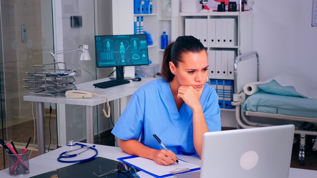 Infirmière médecin fatiguée mettant ses lunettes au repos, les yeux continuaient à regarder l'écran du PC. Médecin en médecine rédige une liste uniforme de patients consultés et diagnostiqués, faisant des recherches.