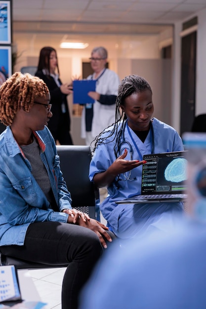 Infirmière et patient analysant l'analyse du cerveau sur un ordinateur portable, parlant du diagnostic de tomographie et du système neuronal sur ordinateur. Jeune femme et assistante médicale faisant une consultation de contrôle dans le hall de la salle d'attente.