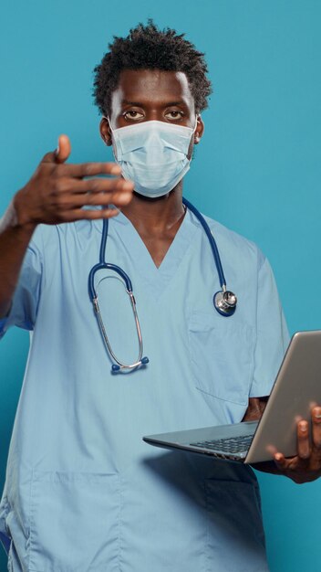 Infirmière portant un masque facial et tenant un ordinateur portable, expliquant l'épidémie de coronavirus. Assistant médical avec uniforme et stéthoscope regardant la caméra tout en ayant un appareil moderne en main