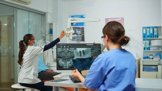 Photo gratuite infirmière stomatologue comparant des radiographies regardant un ordinateur, tandis qu'un médecin spécialiste avec un masque facial parle à un homme souffrant de maux de dents assis sur une chaise stomatologique préparant des outils pour la chirurgie