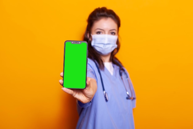 Infirmière tenant un écran vert vertical sur smartphone tout en portant un masque facial contre le coronavirus. Assistant médical montrant un appareil avec clé chroma et modèle de maquette isolé sur l'appareil photo.