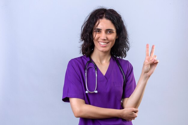 Infirmière en uniforme médical et stéthoscope montrant et pointant vers le haut avec les doigts numéro deux ou signe de la victoire en souriant confiant et heureux sur blanc isolé