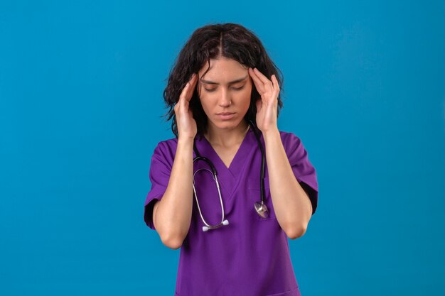 Infirmière en uniforme médical et avec stéthoscope sensation de fatigue touchant les tempes ayant mal de tête debout sur bleu isolé