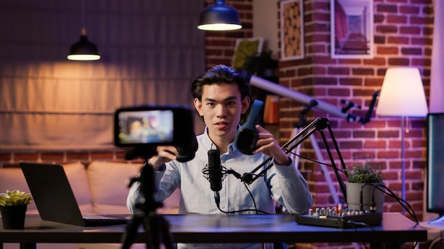 Photo gratuite un influenceur asiatique filmant un examen des écouteurs sur un épisode de podcast, enregistrant une vidéo de recommandation sur caméra. talk-show de diffusion en direct avec casque sans fil sur les réseaux sociaux.