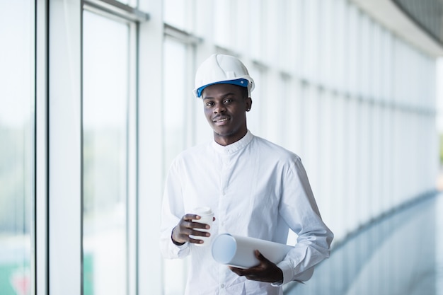 Ingénieur afro-américain de la construction devant le bâtiment tenant des plans contre les fenêtres