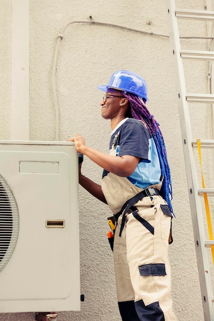 Un ingénieur afro-américain démonte le panneau supérieur en métal du climatiseur extérieur pour vérifier les composants défectueux. Technicien certifié effectuant des réparations sur un système de CVC endommagé