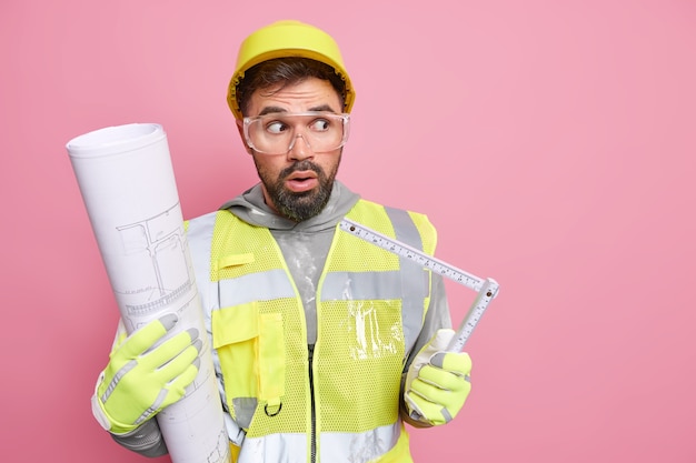 Un ingénieur de chantier de sexe masculin stupéfait travaille avec un plan et un ruban à mesurer prépare un projet de construction vêtu d'un uniforme de casque de sécurité regarde étonnamment loin