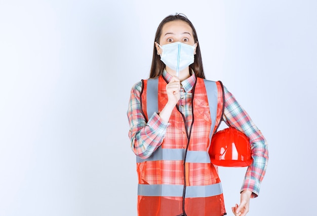 Ingénieur en construction féminin dans un masque de sécurité et un casque rouge sous ses bras tenant un stylo et a l'air confus et réfléchi