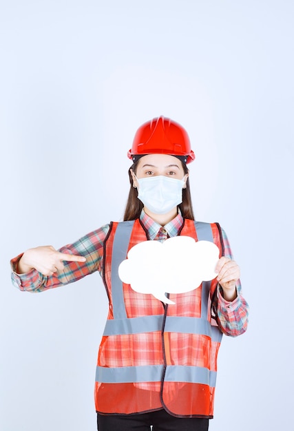 Ingénieur en construction féminin en masque de sécurité et casque rouge tenant un tableau d'informations en forme de nuage.