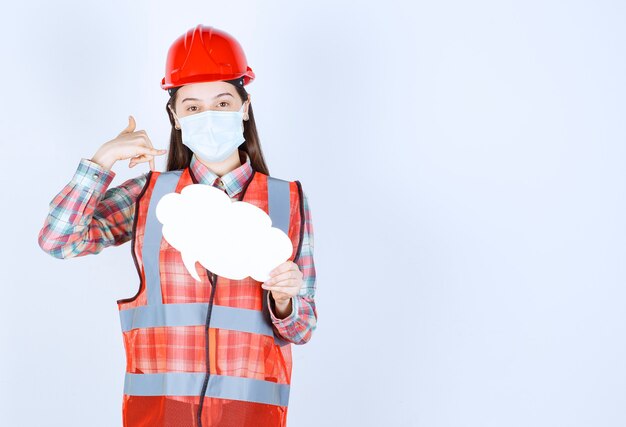 Ingénieur en construction féminin portant un masque de sécurité et un casque rouge tenant un panneau d'information en forme de nuage et demandant un appel