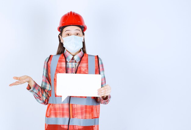 Ingénieur en construction féminin portant un masque de sécurité et un casque rouge tenant un panneau d'information vierge rectangulaire et ayant l'air confus et réfléchi