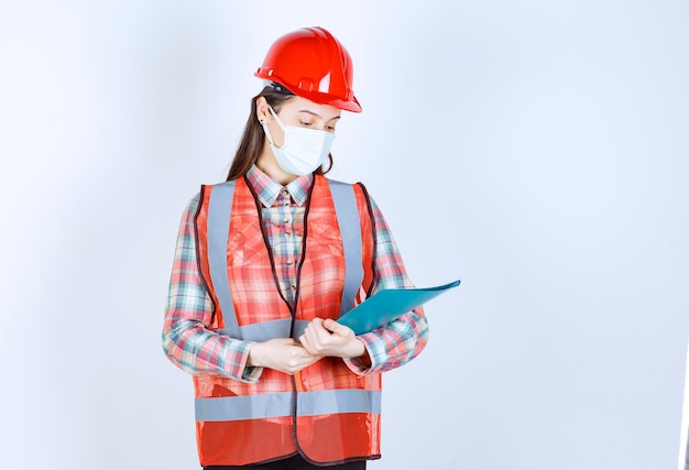 Ingénieur en construction en masque de sécurité et casque rouge tenant un dossier bleu.