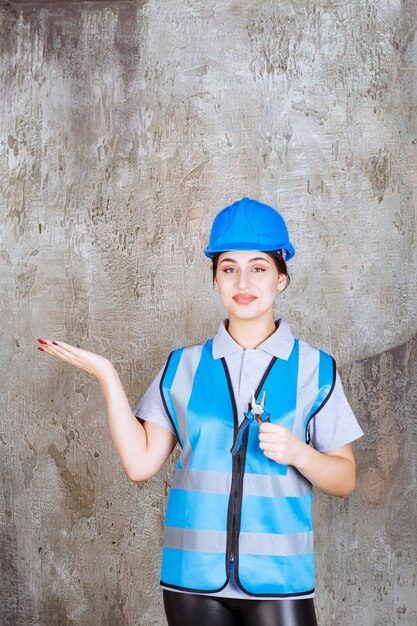 Ingénieur féminin en uniforme bleu et casque tenant des pinces métalliques pour réparation et montrant le mur de béton derrière