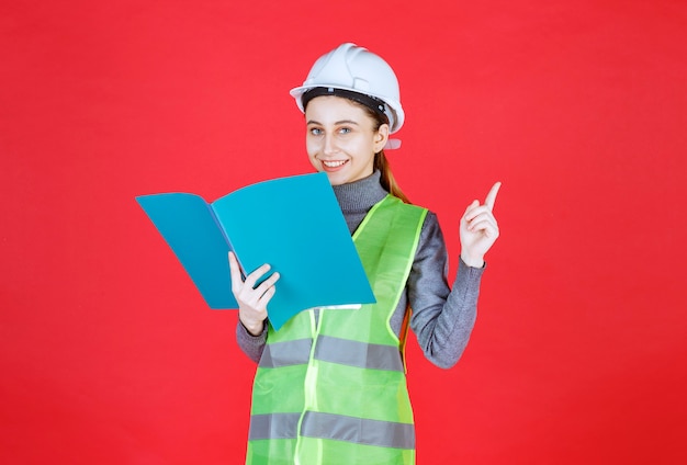 Ingénieur femme avec casque blanc tenant un fichier de projet bleu, le lisant et réfléchissant.