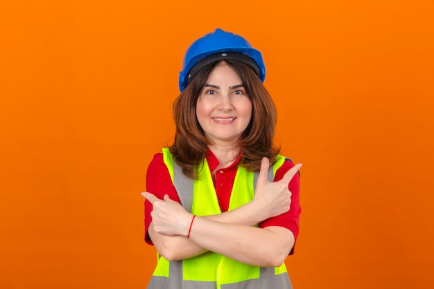 Ingénieur femme portant un gilet de construction et un casque de sécurité debout avec les bras croisés pointant avec l'index vers d'autres côtés avec grand sourire sur le visage sur zone orange isolée