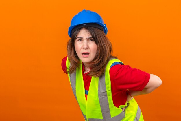 Ingénieur femme portant un gilet de construction et un casque de sécurité à la recherche de maux de dos malaise debout sur mur orange isolé