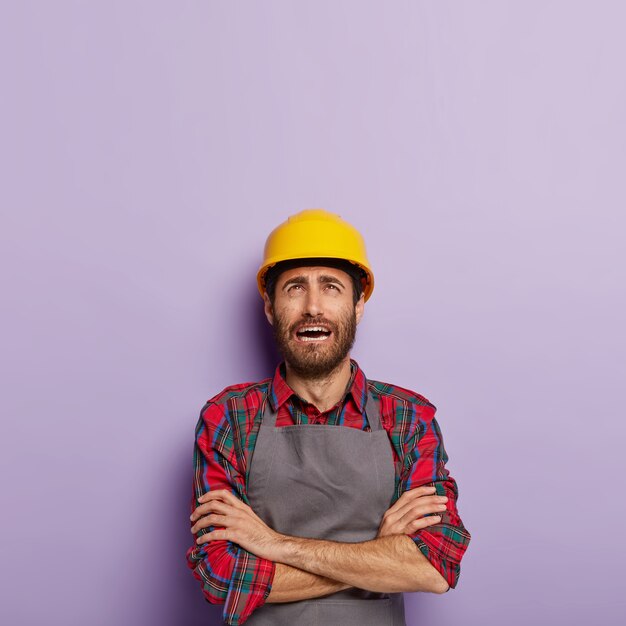 L'ingénieur masculin insatisfait stressant garde les bras croisés a une expression de visage misérable, porte une chemise à carreaux et un tablier, concentré dessus, obtient beaucoup de tâches du patron fatigué du travail manuel isolé sur violet