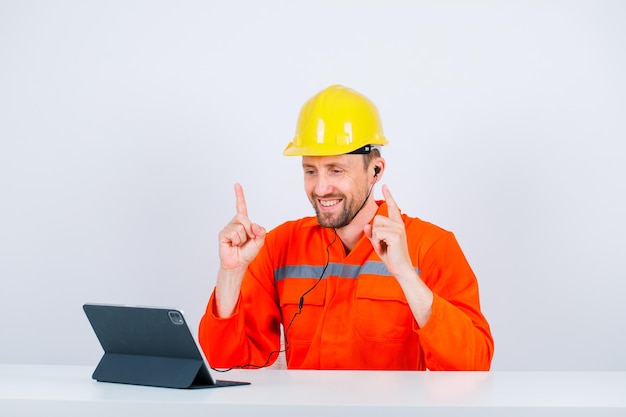 Ingénieur souriant pointe vers le haut avec les index en s'asseyant devant sa tablette sur fond blanc