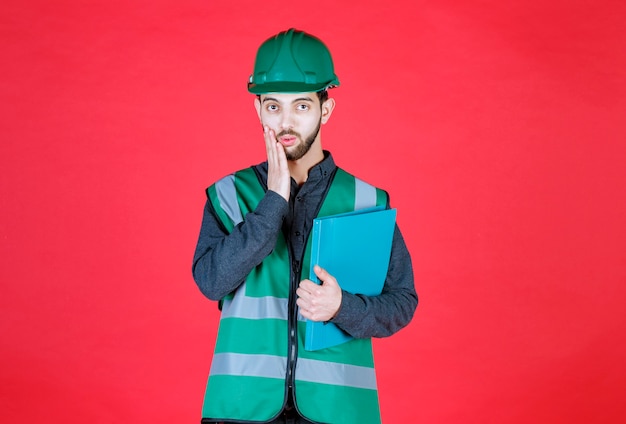 Ingénieur en uniforme vert et casque tenant un dossier bleu, a l'air terrifié et ravi.