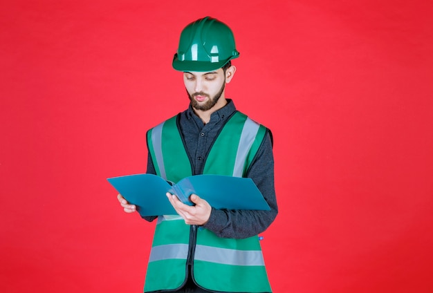 Ingénieur en uniforme vert et casque tenant un dossier bleu, l'ouvrant et le lisant.