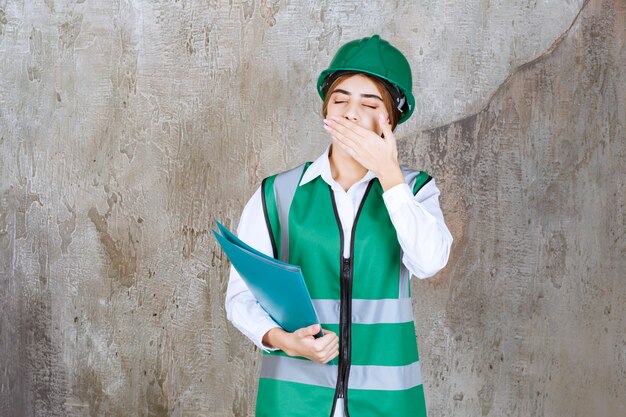 Ingénieur en uniforme vert et casque tenant un dossier de projet vert et a l'air fatigué et somnolent.