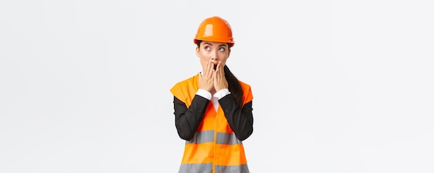 Une ingénieure asiatique choquée et inquiète dans un casque de sécurité et une veste réfléchissante regardant dans le coin supérieur gauche et haletant un regard inquiet étonné debout sur fond blanc