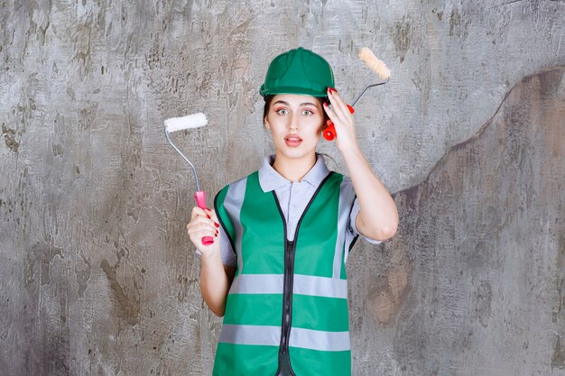 Ingénieure en casque vert tenant un rouleau de finition pour la peinture murale et réfléchissant à de nouvelles méthodes