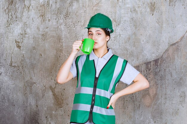 Ingénieure en casque vert tenant une tasse de café vert et buvant.