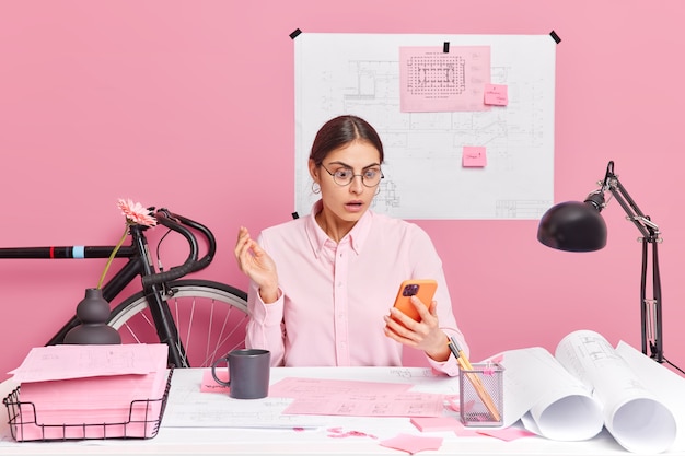 Une ingénieure professionnelle qualifiée concentrée sur l'affichage d'un smartphone avec une expression choquée fait des dessins et des plans de pose sur le bureau