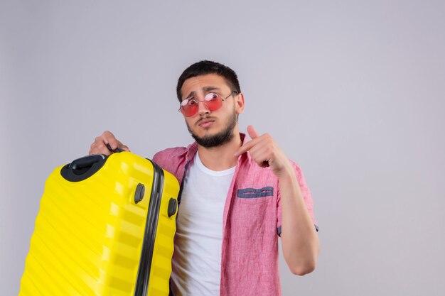 Inquiet jeune homme beau voyageur portant des lunettes de soleil regardant la caméra avec une expression de confusion pointant avec le doigt sur sa valise debout sur fond blanc