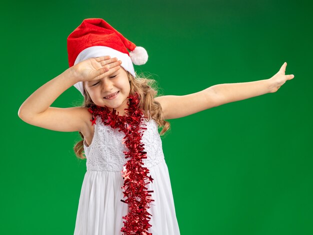 Insatisfait aux yeux fermés petite fille portant un chapeau de Noël avec guirlande sur le cou mettant la main sur le front et les points sur le côté isolé sur fond vert avec espace copie