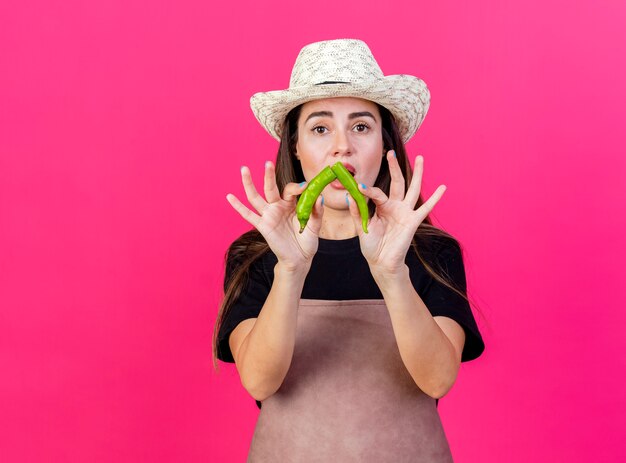 Insatisfait belle fille de jardinier en uniforme portant chapeau de jardinage a cassé le poivre isolé sur rose