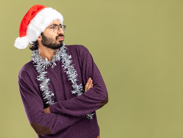 Insatisfait à côté jeune beau mec portant un chapeau de Noël avec guirlande sur le cou croisant les mains isolés sur fond vert olive avec espace copie