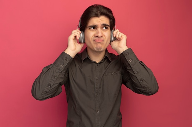 Insatisfait à côté jeune beau mec portant un t-shirt noir avec un casque isolé sur un mur rose