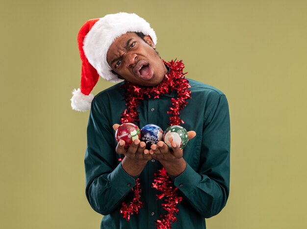 Insatisfait homme afro-américain en bonnet de noel avec guirlande tenant des boules de Noël regardant la caméra avec une expression de confusion debout sur fond vert