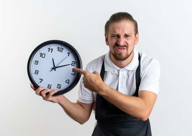 Insatisfait jeune beau coiffeur en uniforme tenant et pointant sur l'horloge isolé sur fond blanc