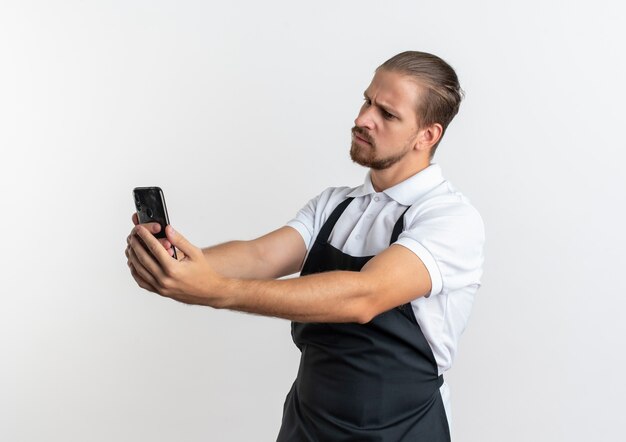 Insatisfait jeune beau coiffeur en uniforme tenant et regardant un téléphone mobile isolé sur fond blanc