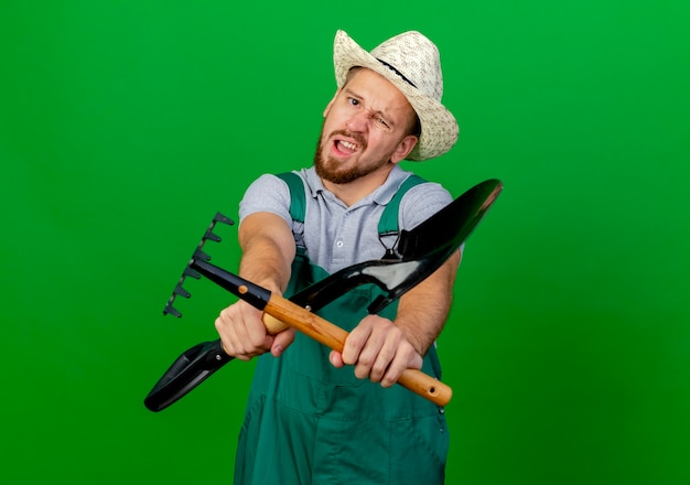 Insatisfait jeune beau jardinier slave en uniforme et chapeau à la pelle et râteau vers